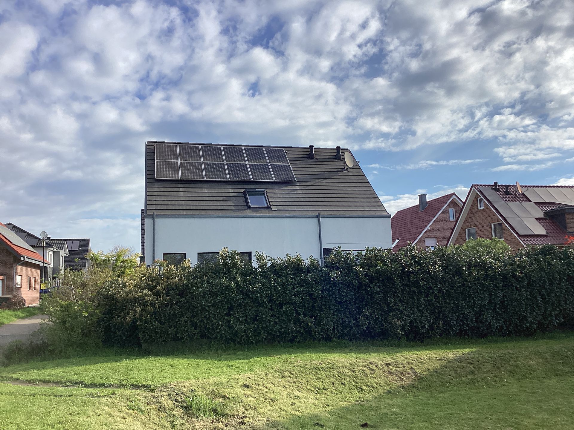 Haus mit Solarpaneelen auf dem Dach, umgeben von grünen Sträuchern, unter bewölktem Himmel.