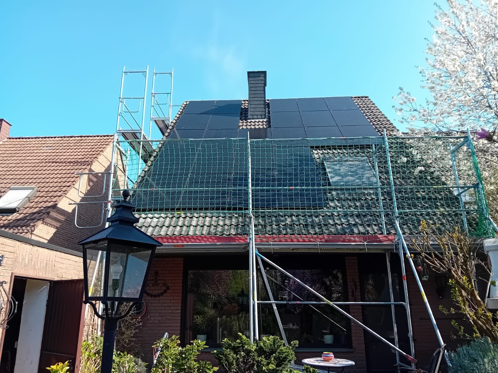 Auf einem Dach mit Gerüst, Schornstein und strahlend blauem Himmel werden Solarpaneele installiert.