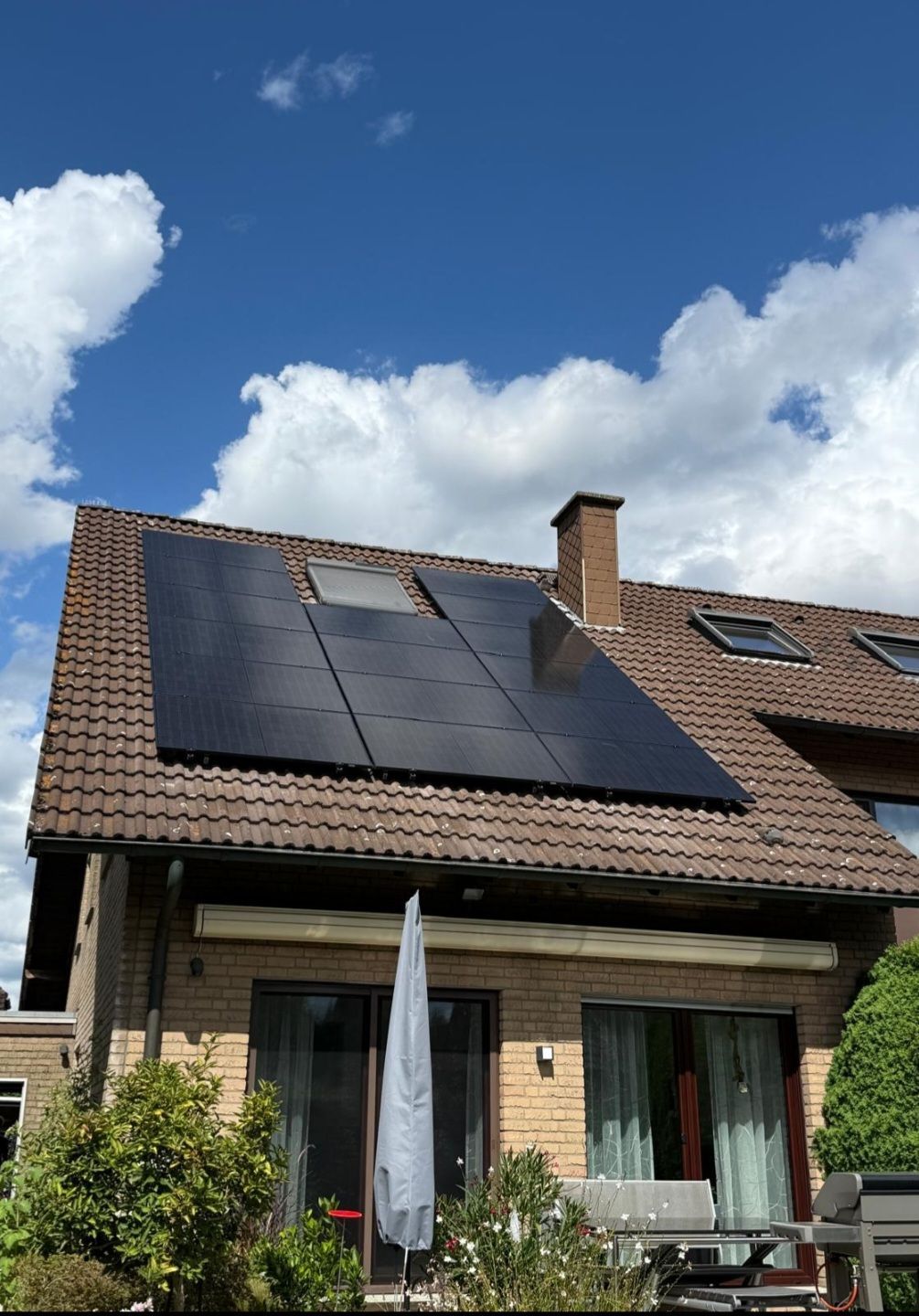 Solarpaneele auf einem Ziegeldach unter leicht bewölktem, blauem Himmel. Backsteinhaus, Terrassenmöbel.