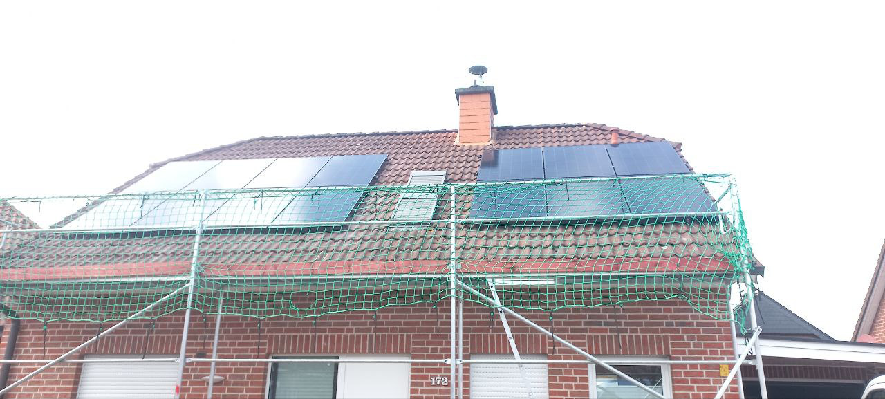 Haus mit Solarpaneelen auf einem roten Ziegeldach, umgeben von Gerüsten und grünen Netzen, unter einem bewölkten Himmel.