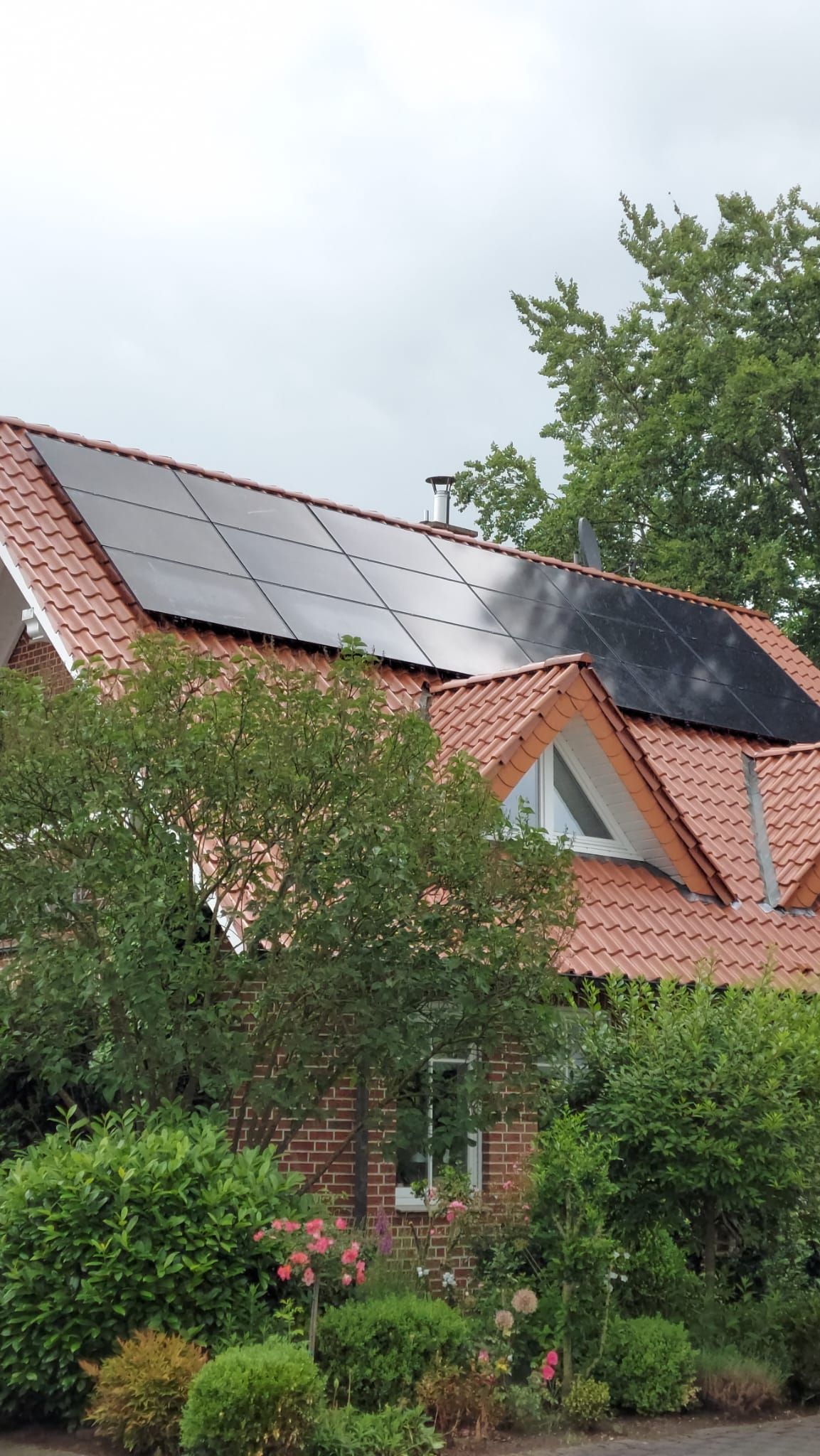 Haus mit rotem Ziegeldach, Solarpaneelen und umgebender Vegetation unter bedecktem Himmel.