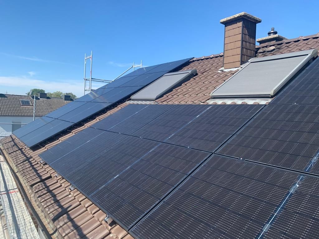 Solarpaneele auf einem Ziegeldach mit Oberlichtern und einem Schornstein vor blauem Himmel.