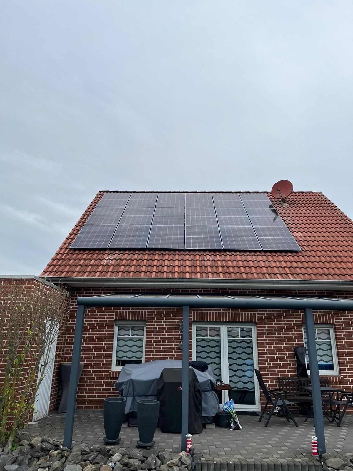Solarpaneele auf einem roten Ziegeldach über einem Backsteinhaus mit Terrasse und bedecktem Himmel.