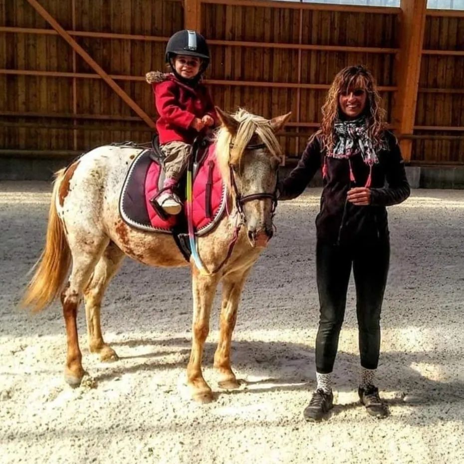 Un enfant monte un petit cheval à l'intérieur, à côté d'un moniteur, tous deux portant une tenue d'équitation.