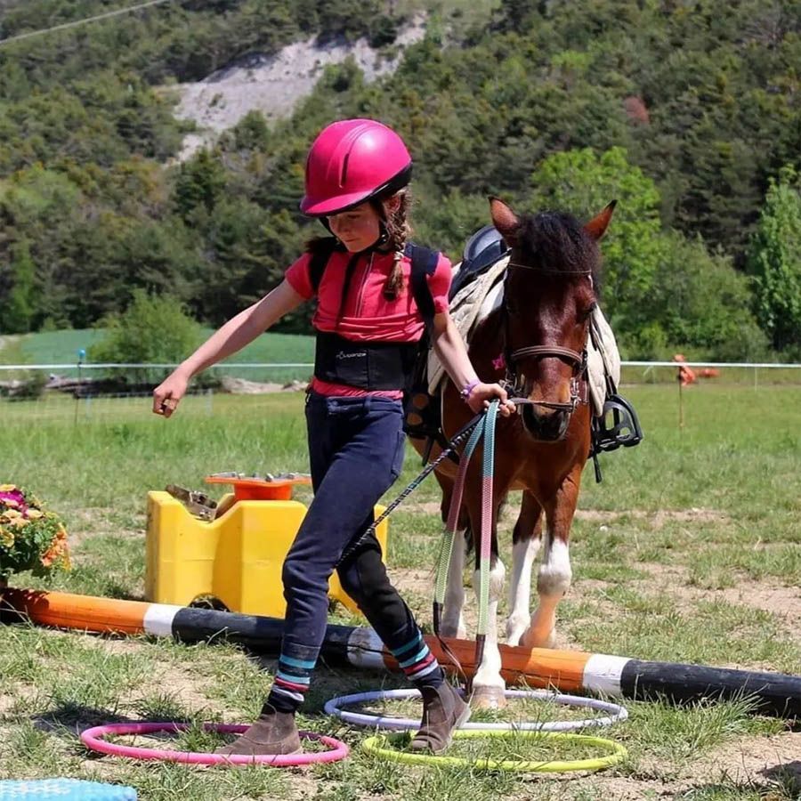 Enfant portant un casque rose menant un cheval à travers des anneaux colorés sur un parcours équestre extérieur herbeux