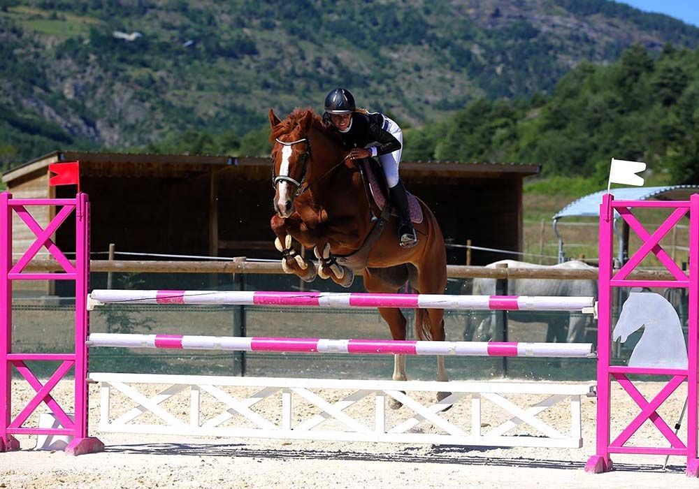 Un cheval et son cavalier franchissent un obstacle rose et blanc dans un manège équestre extérieur.