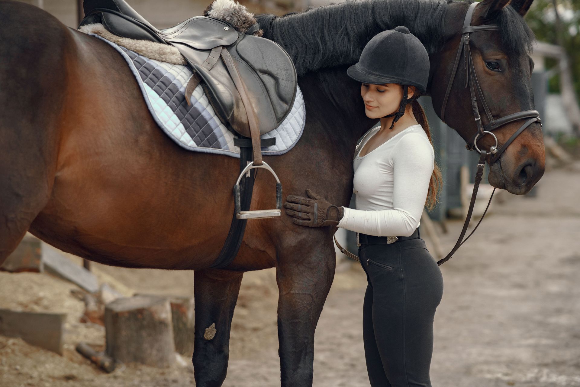 Une femme portant un casque d'équitation se tient à côté d'un cheval sellé à l'extérieur.