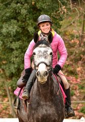 Cavalier souriant en veste rose sur un cheval noir, en extérieur sur un sentier boisé