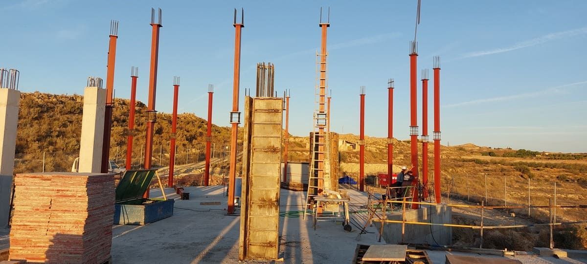Obra en construcción con pilares de hormigón y vigas de acero rojas; al aire libre con luz natural.