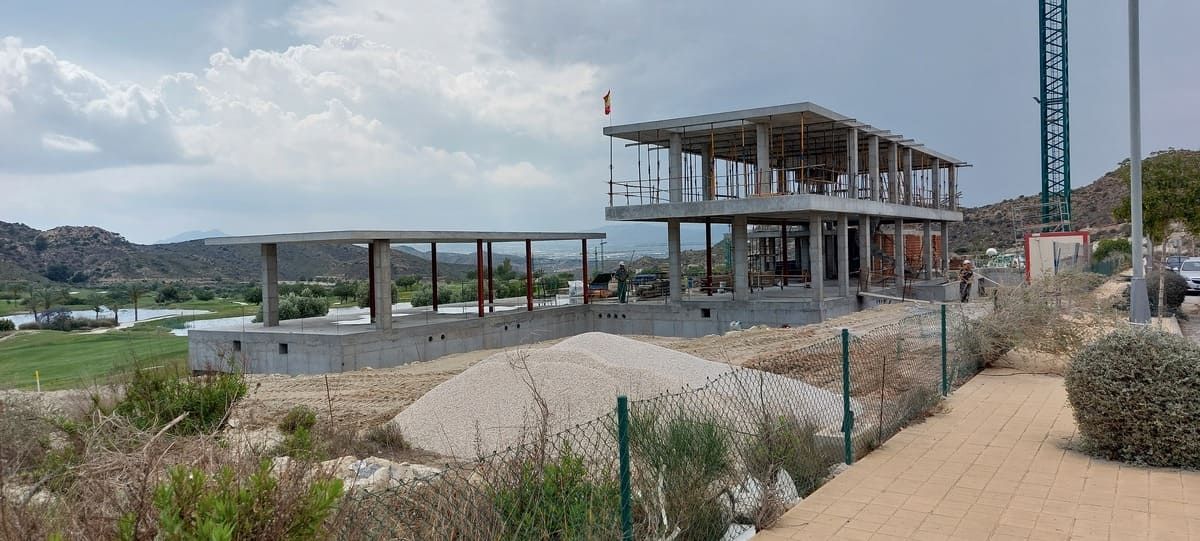 Sitio de construcción de un edificio de dos pisos con estructura de concreto sin terminar, rodeado de tierra, vegetación y una grúa.