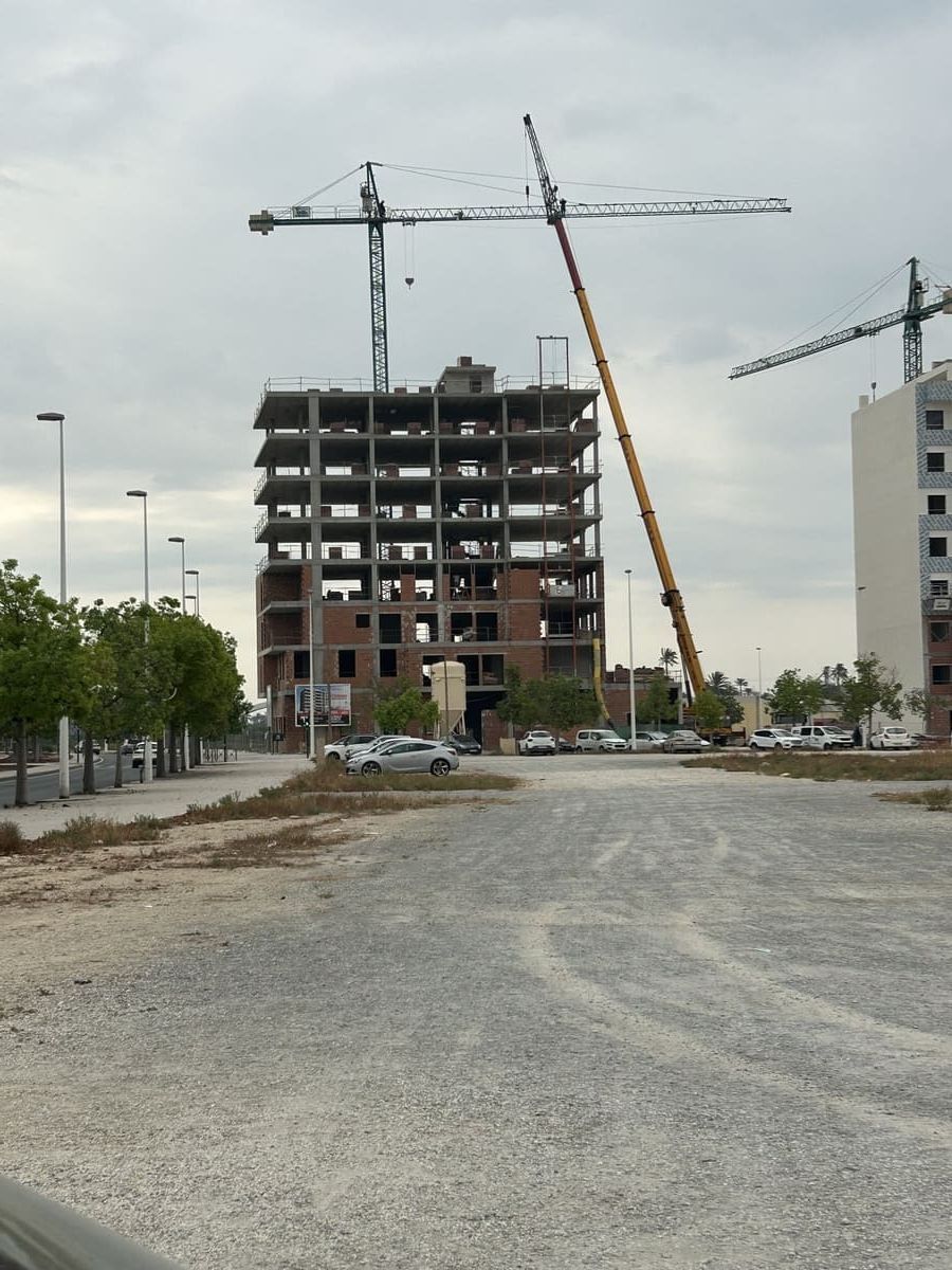 Un edificio de varios pisos en construcción con dos grúas en un lote de tierra bajo un cielo nublado.