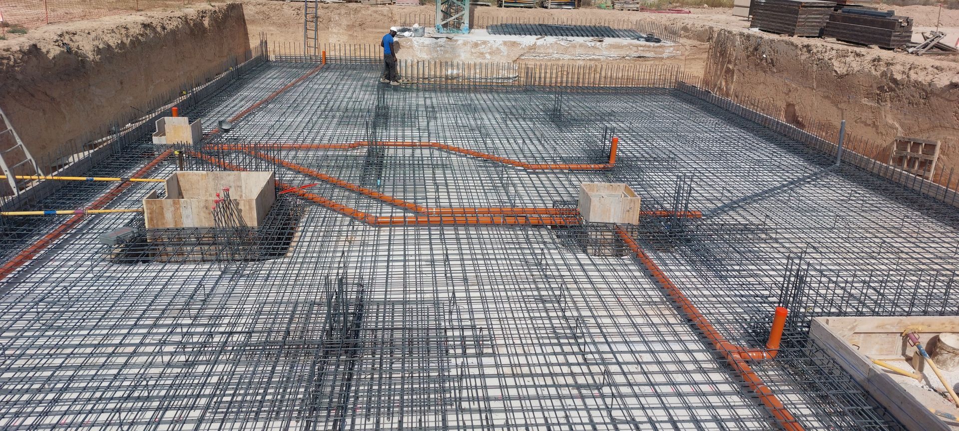 Sitio de construcción con rejilla de varilla de refuerzo, conductos y trabajadores colocando los cimientos.