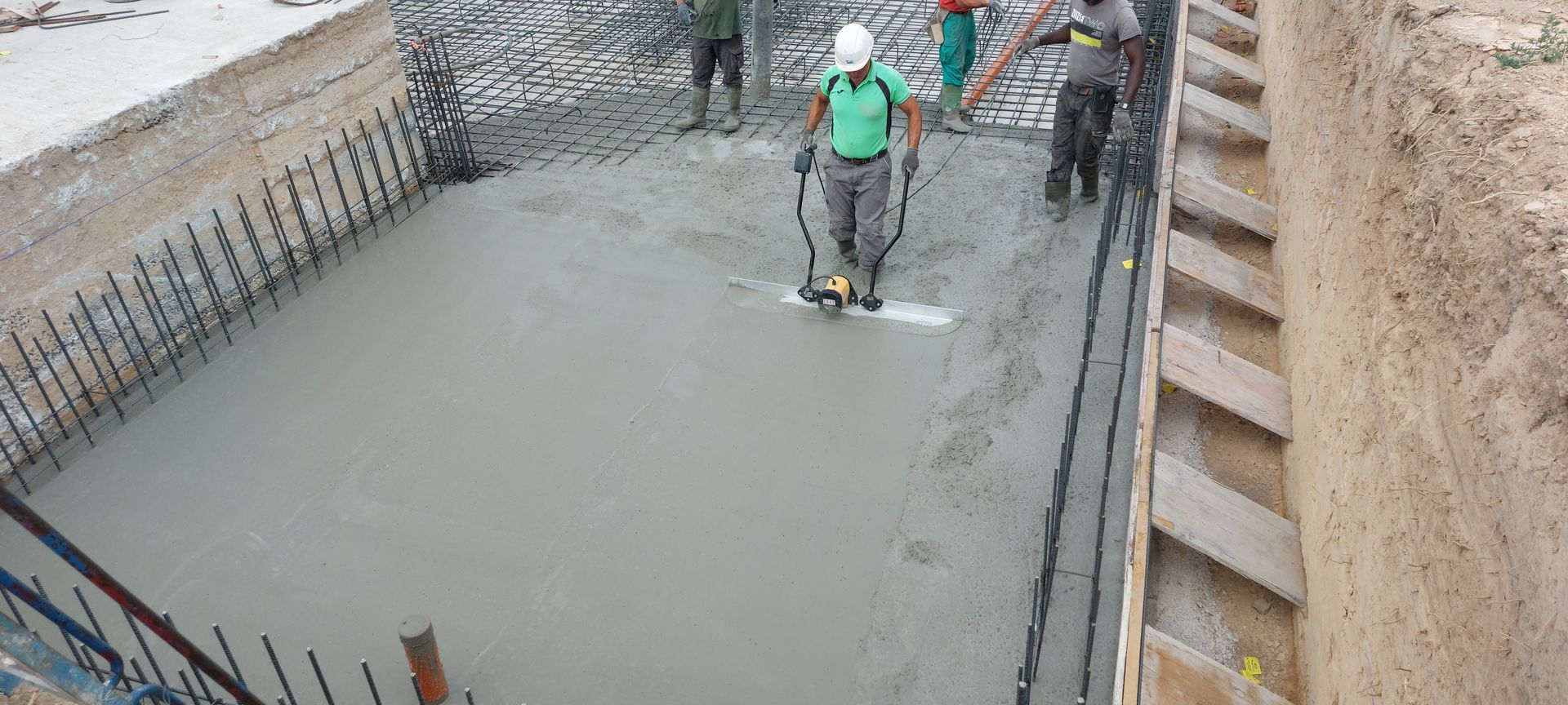 Trabajadores alisan hormigón fresco en un sitio de construcción con barras de refuerzo y estructura de madera.