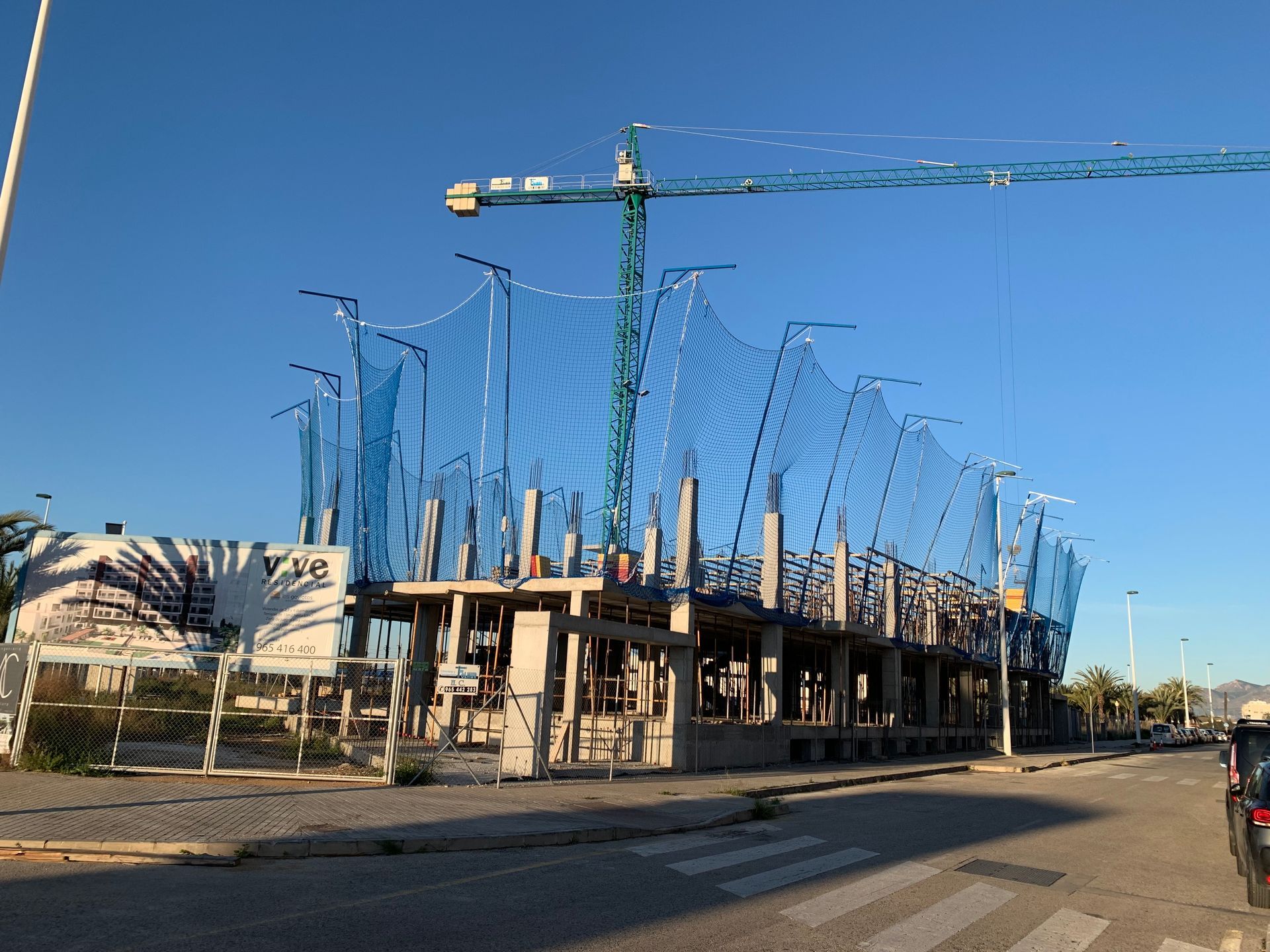 Sitio de construcción con red de seguridad azul, grúa verde y un cielo azul claro.