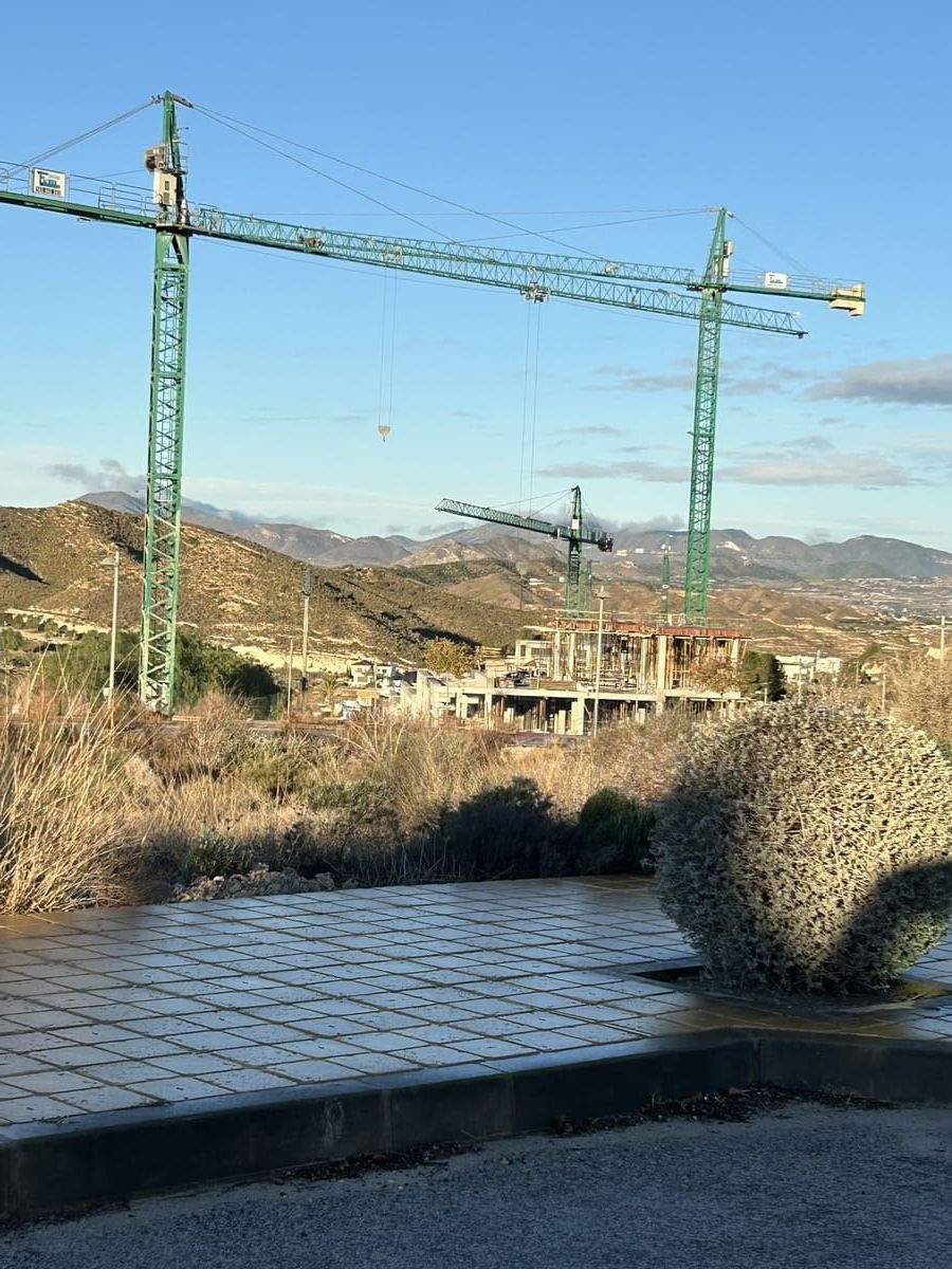 Dos grúas de construcción verdes sobre una obra en construcción, con colinas al fondo bajo un cielo soleado.