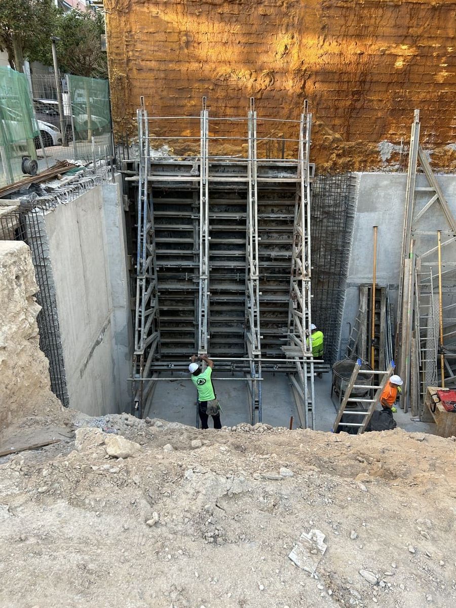 Obreros de la construcción en una zanja con andamios contra un muro de ladrillo, con paredes de hormigón. Tierra en primer plano.