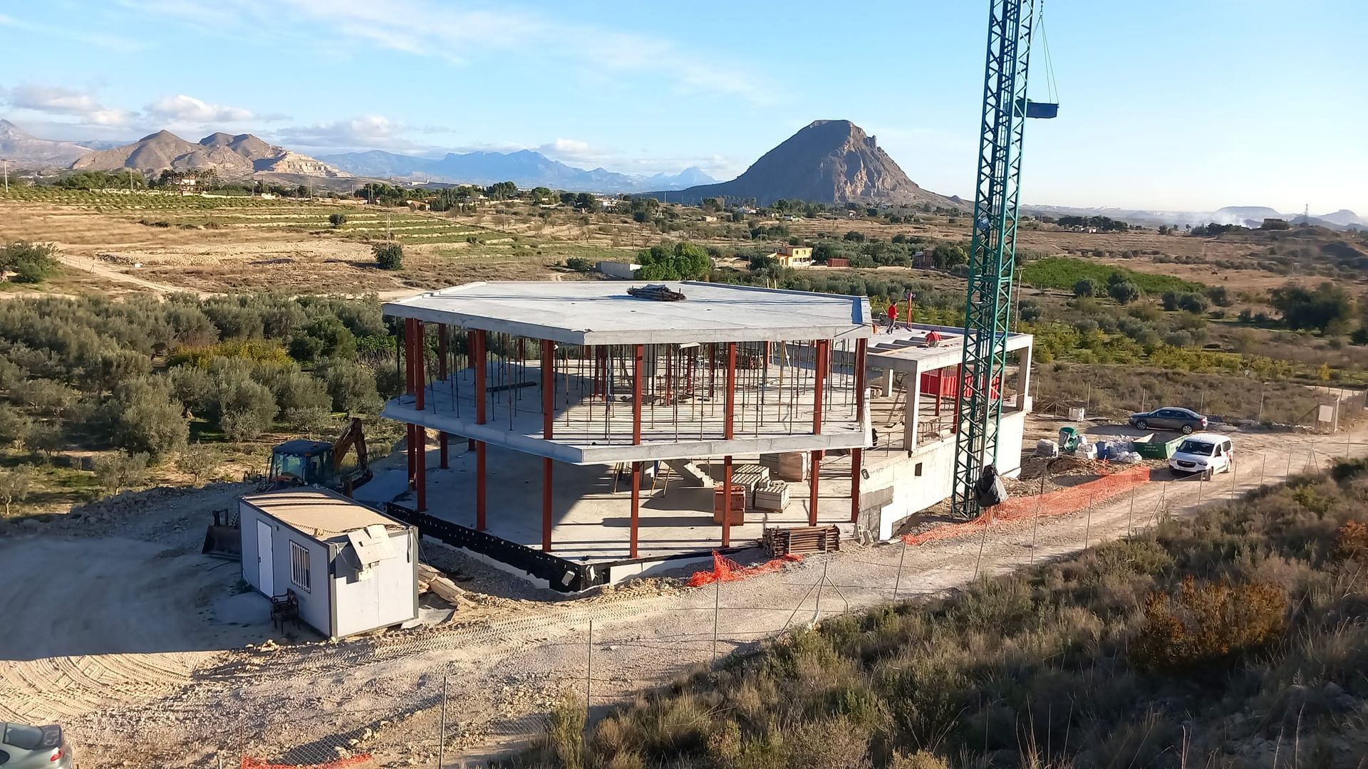 Sitio de construcción de un edificio circular de dos pisos con grúa en un paisaje rural, cielo azul.