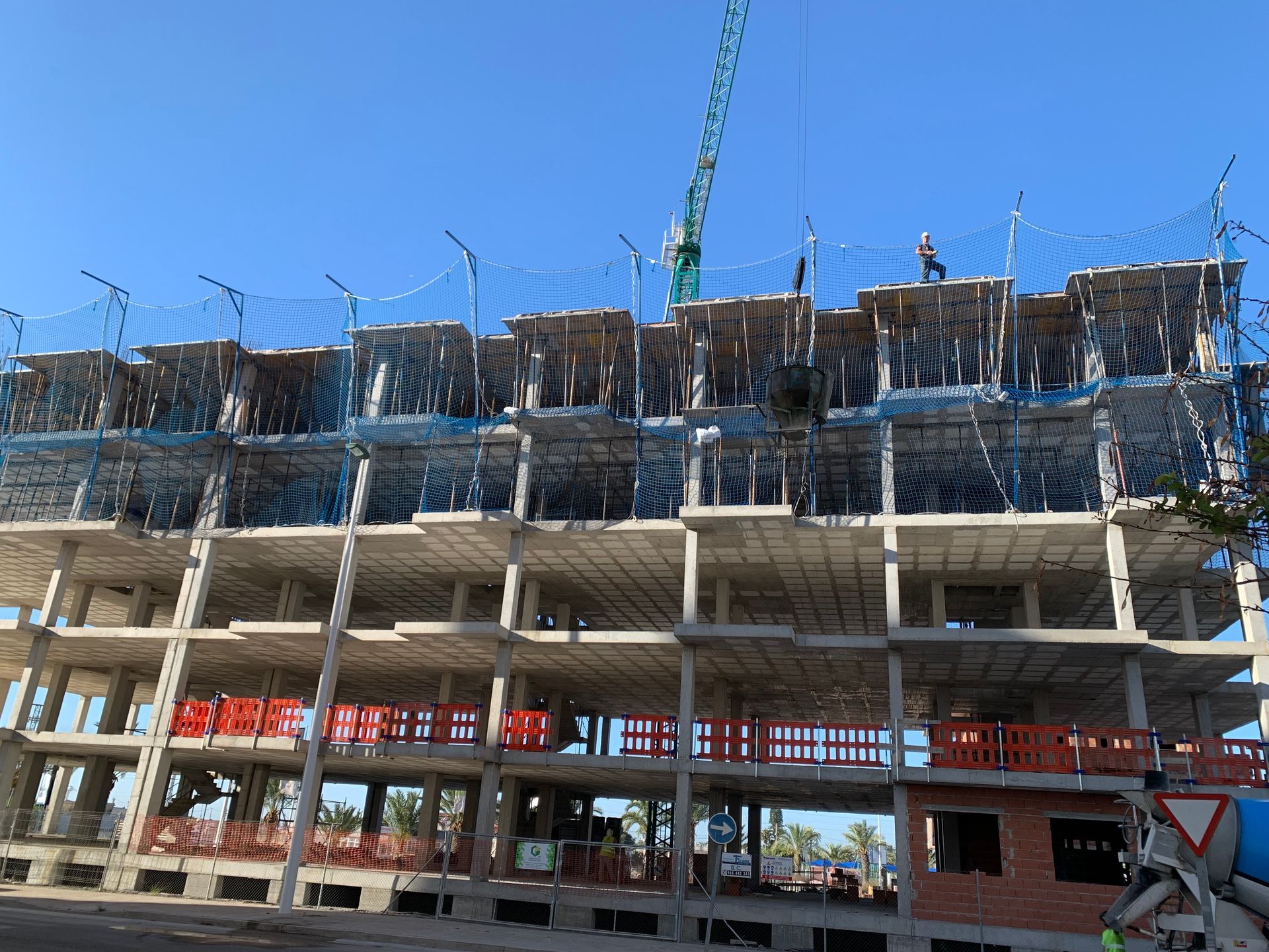 Edificio de hormigón de varios pisos en construcción con red de seguridad, cielo azul, grúa visible.
