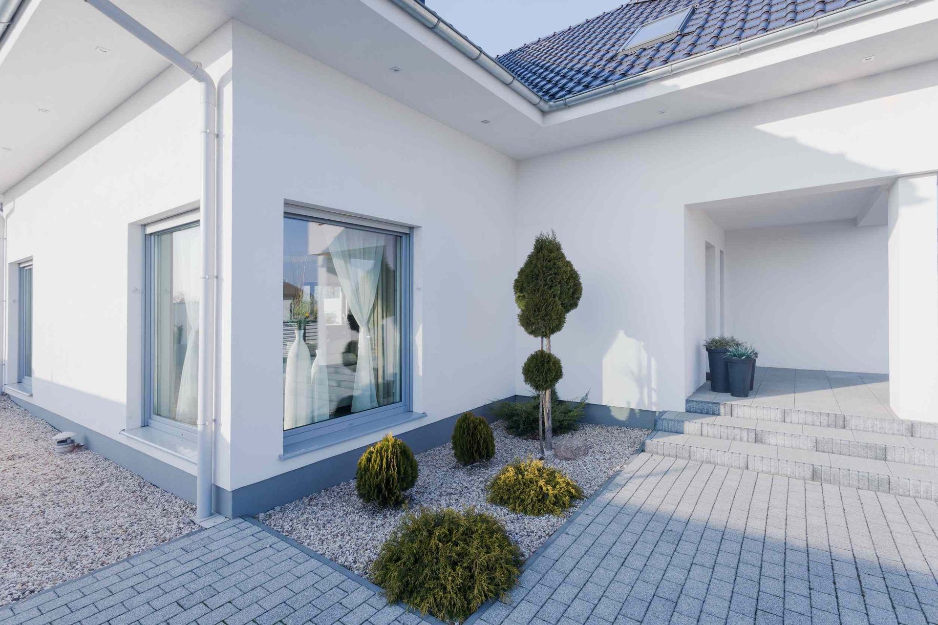 Exterior de casa moderna blanca con entrada ajardinada, patio de ladrillo gris y pequeños arbustos de hoja perenne.