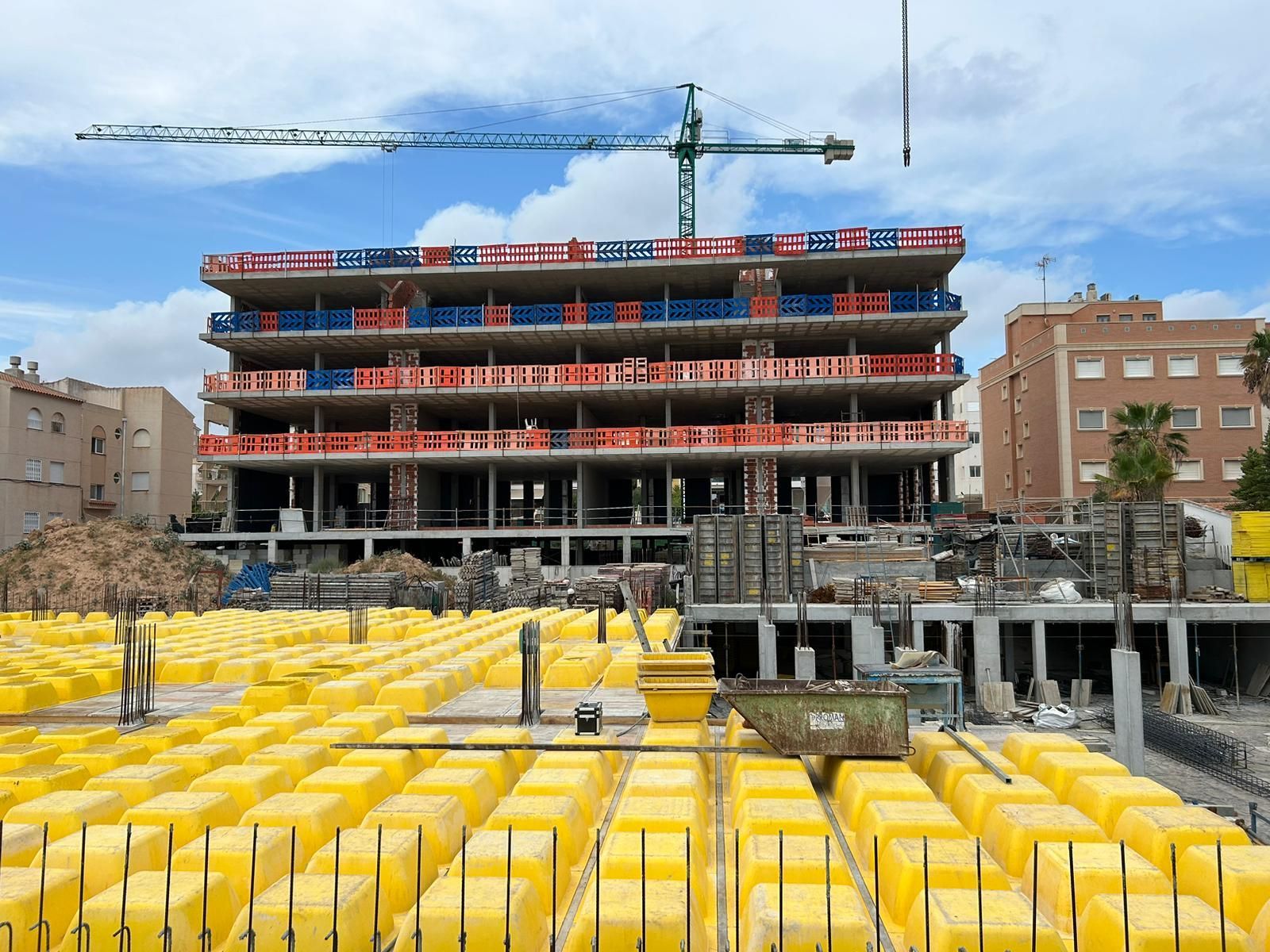 Sitio de construcción de un edificio de varios pisos, bloques amarillos en primer plano, grúa arriba, día soleado.