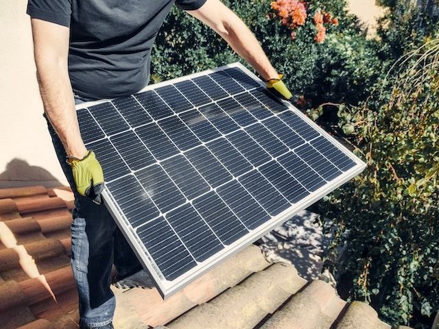 Un hombre sostiene un panel solar en la parte superior de un tejado.