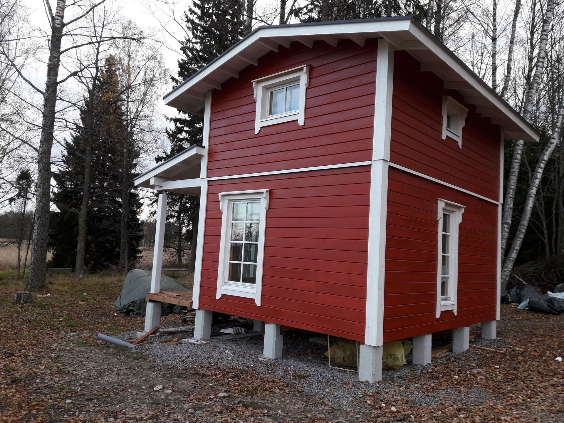 Pieni, kaksikerroksinen punainen puinen mökki, jossa on valkoiset reunat, seisoo betonipilareiden päällä metsäisellä alueella, joka on peittynyt pudonneisiin lehtiin.