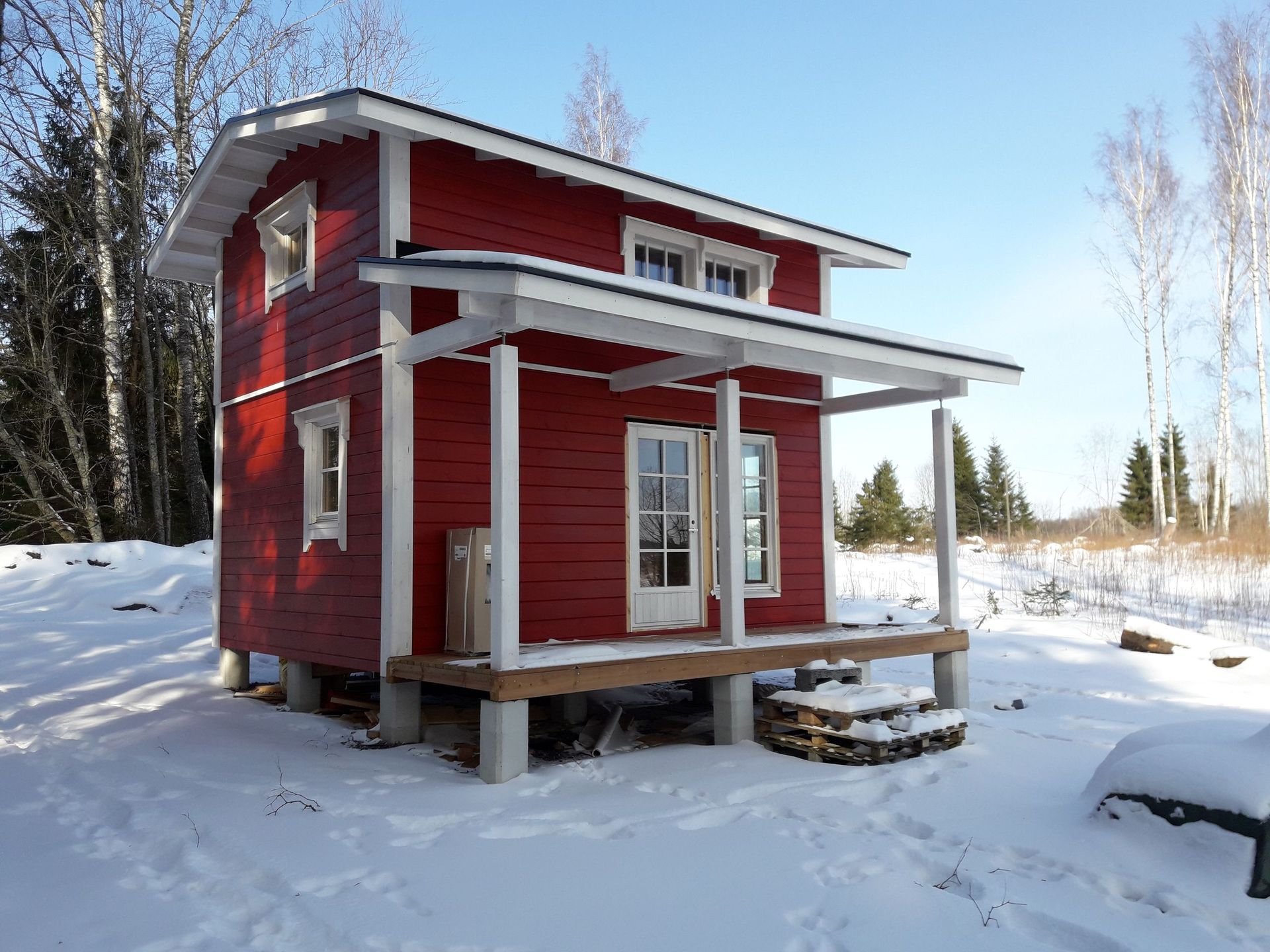 Pieni, kaksikerroksinen punainen puinen mökki kuistineen seisoo lumisissa, metsäisissä maisemissa kirkkaana, aurinkoisena päivänä.