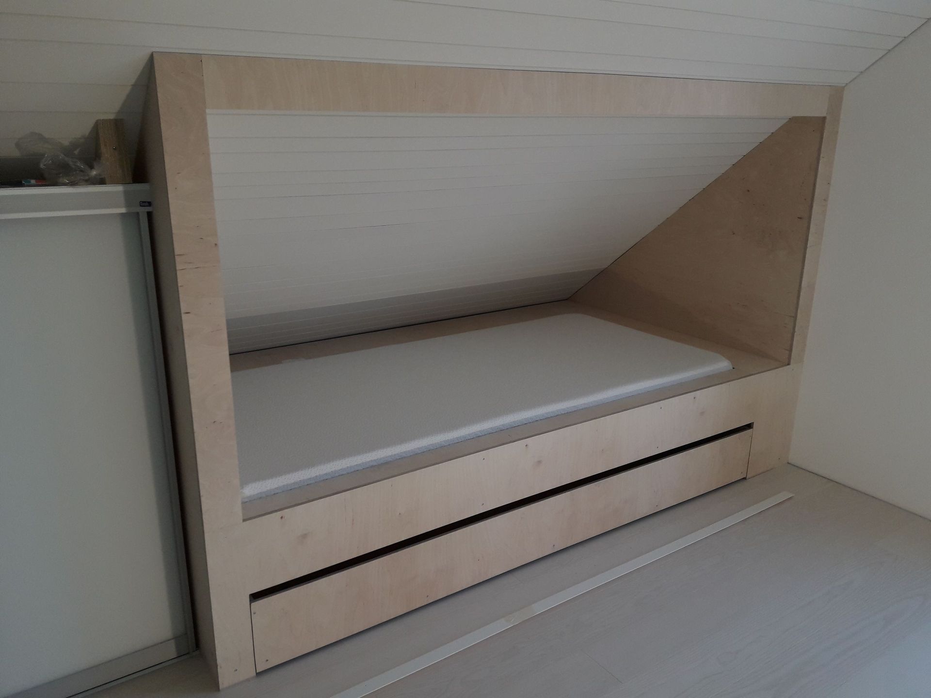 Mittatilaustyönä valmistettu puinen sängynrunko, jossa on sisäänrakennettu laatikko, viistokattoisessa ullakkotilassa.