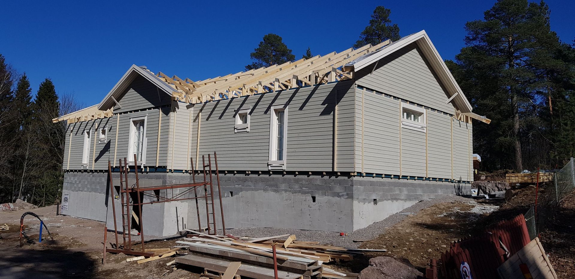 Rakennustyömaa, jossa näkyy yksikerroksinen talo, jossa on vaalea ulkoverhous ja betoninen perustus, kirkkaan sinisen taivaan alla.