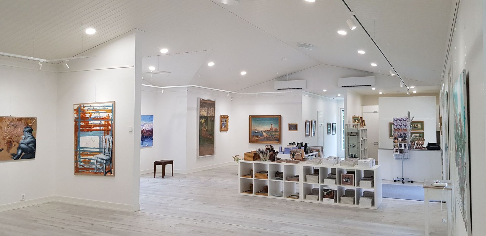 Taidegalleria, jossa on erilaisia ​​kehystettyjä maalauksia valkoisilla seinillä ja keskeinen säilytysyksikkö hyllyineen.