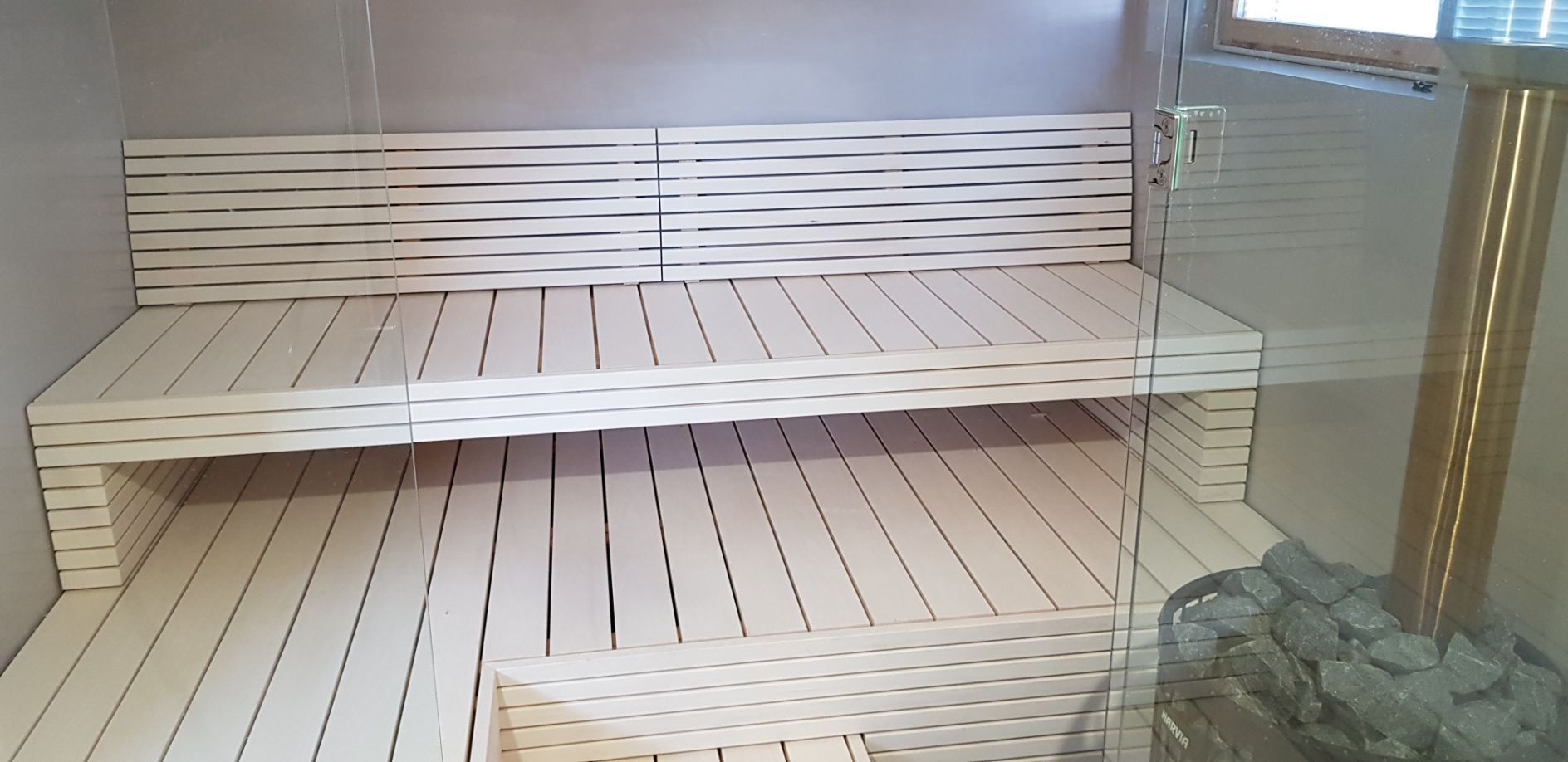 Saunan sisustus, jossa on vaaleat puusälelauteet ja lasilevyn takana oleva kivillä täytetty kiuas.