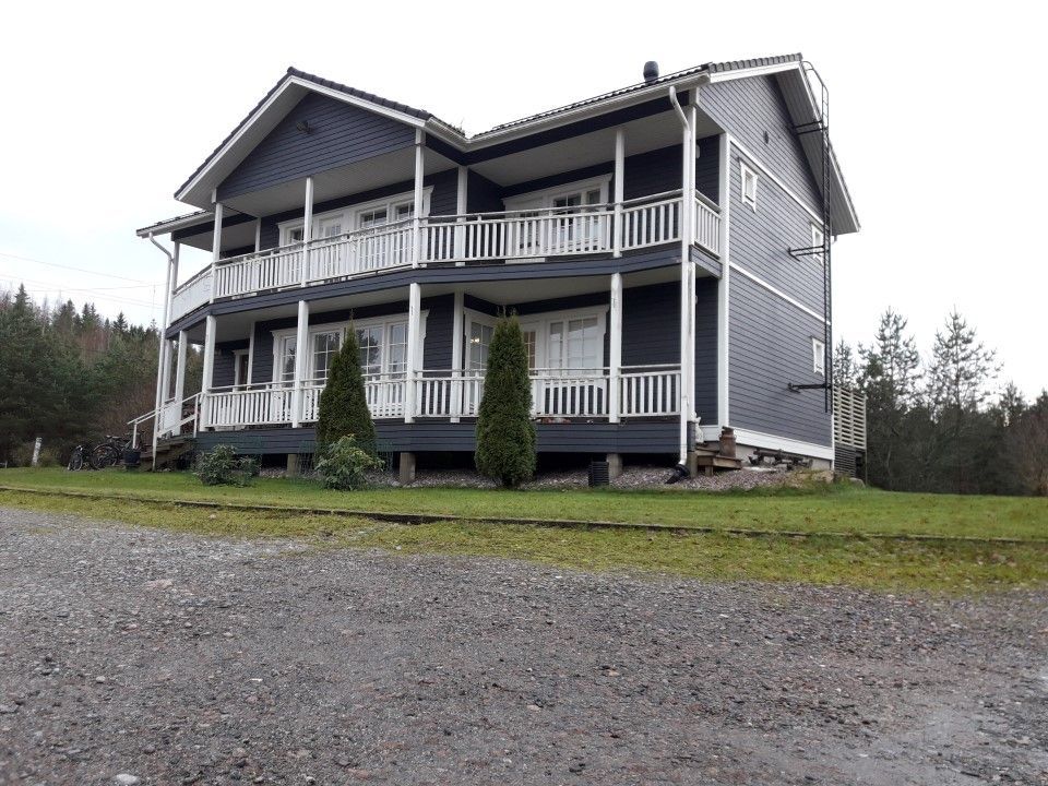 Kaksikerroksinen tummanharmaa puutalo, jossa on valkoiset kaiteet, taloa kiertävät parvekkeet ja pienet männyt etupihalla.