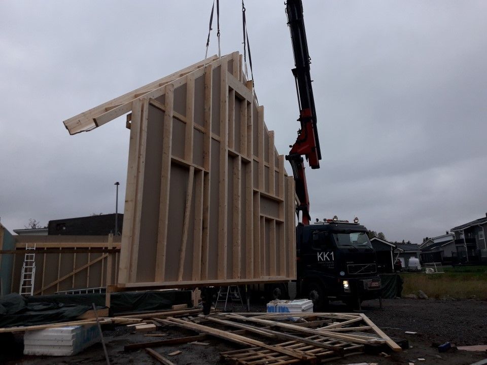 Nosturi nostaa suurta, puista elementtitalon seinäpaneelia rakennustyömaalla pilvisenä päivänä.