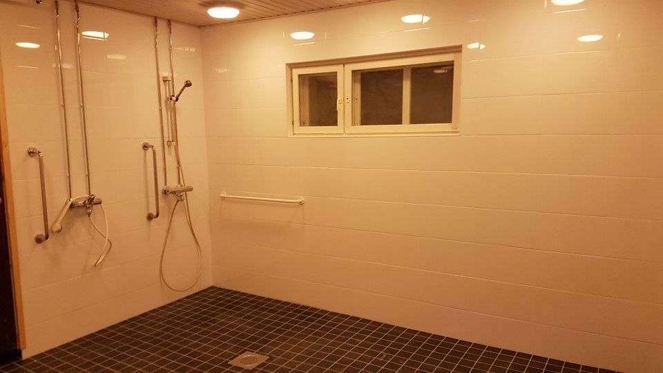 Suihkuhuone, jossa on kaksi suihkupäätä, kaiteet, valkoiset laattaseinät ja tummat neliönmuotoiset lattialaatat.