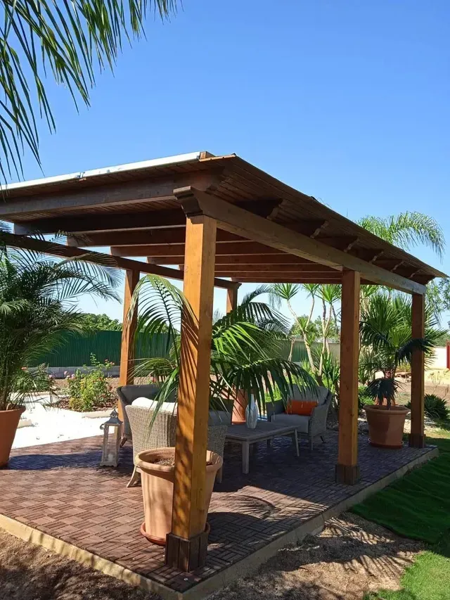 Pérgola de madera sobre un patio de ladrillo con plantas en macetas y asientos bajo un cielo azul.