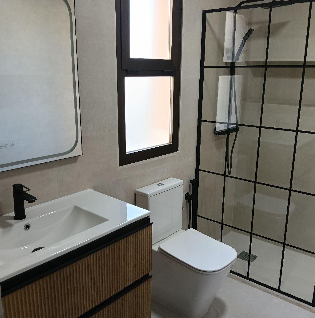 Baño con inodoro, lavabo y ducha de color blanco, con ventana y mampara metálica de color negro.