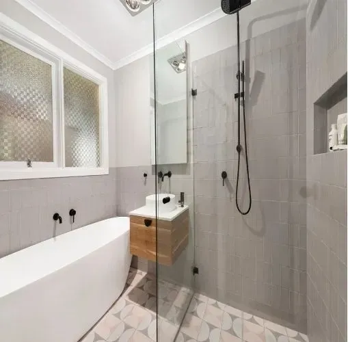 Baño moderno con azulejos grises, bañera independiente, ducha de cristal y tocador de madera.