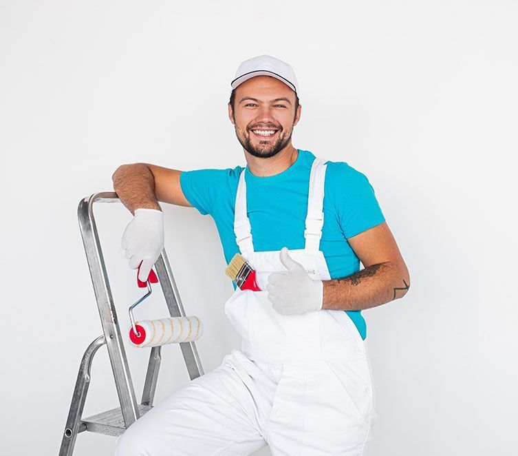Ein Mann sitzt mit einer Farbrolle und einem Pinsel in der Hand auf einer Leiter.