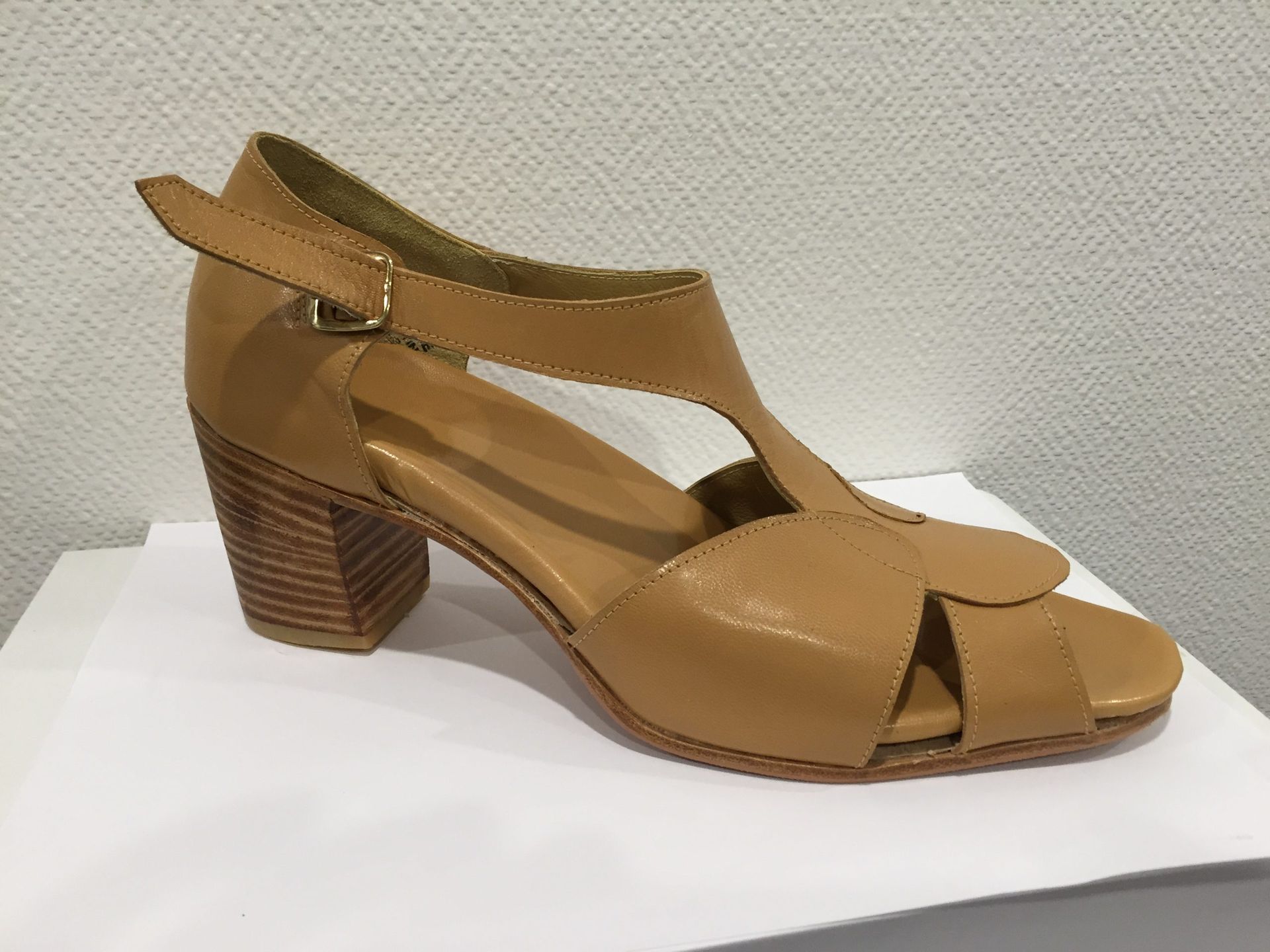 Sandale à talon en cuir beige avec bride à la cheville, présentée sur une surface blanche devant un mur uni.