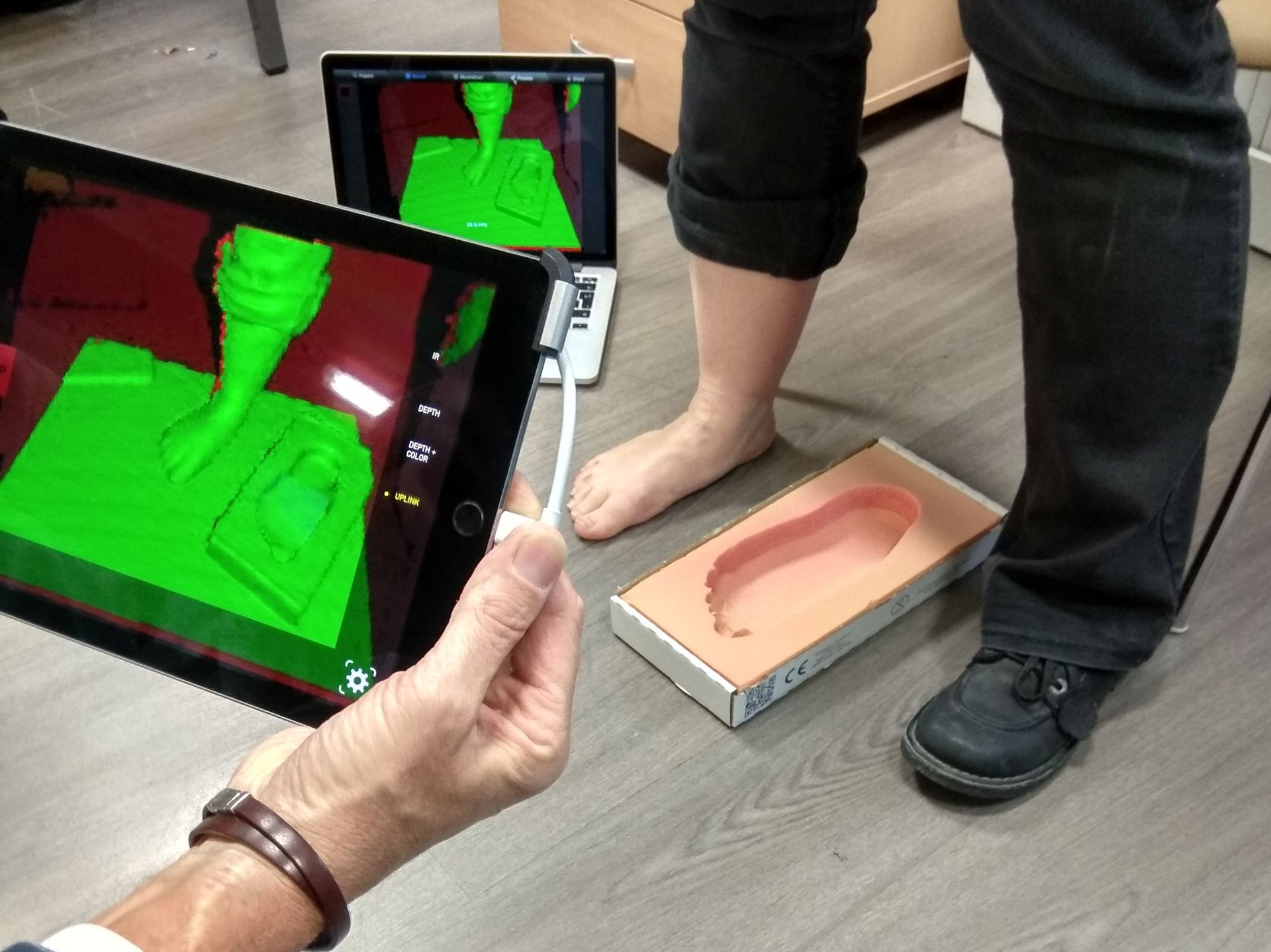 La tablette affiche un scan 3D du pied tandis qu'une personne se tient à côté d'un moule en mousse posé au sol.