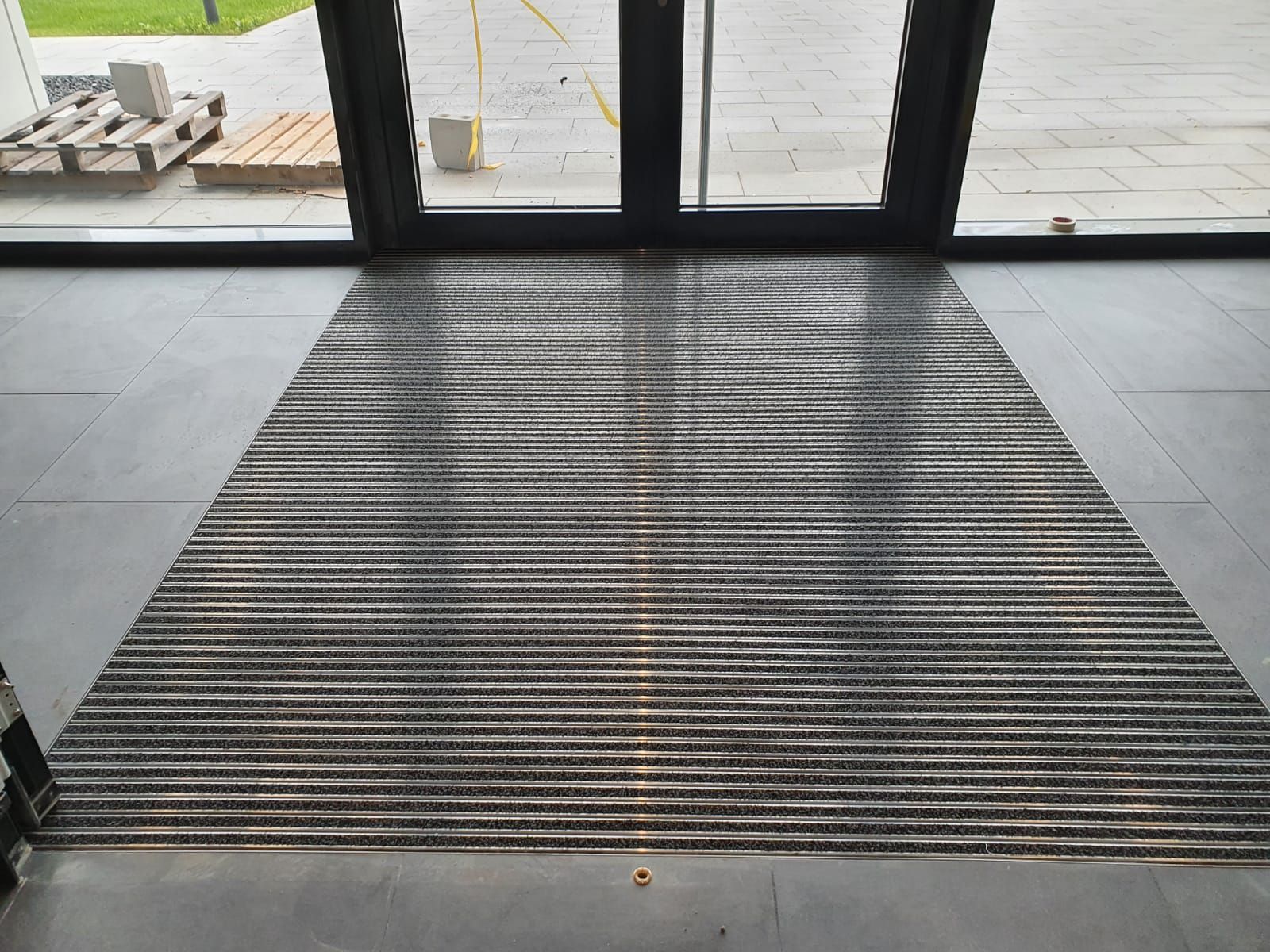 Eingangsbereich mit grauer Fußmatte und Glastür, draußen Paletten und Pflastersteine sichtbar