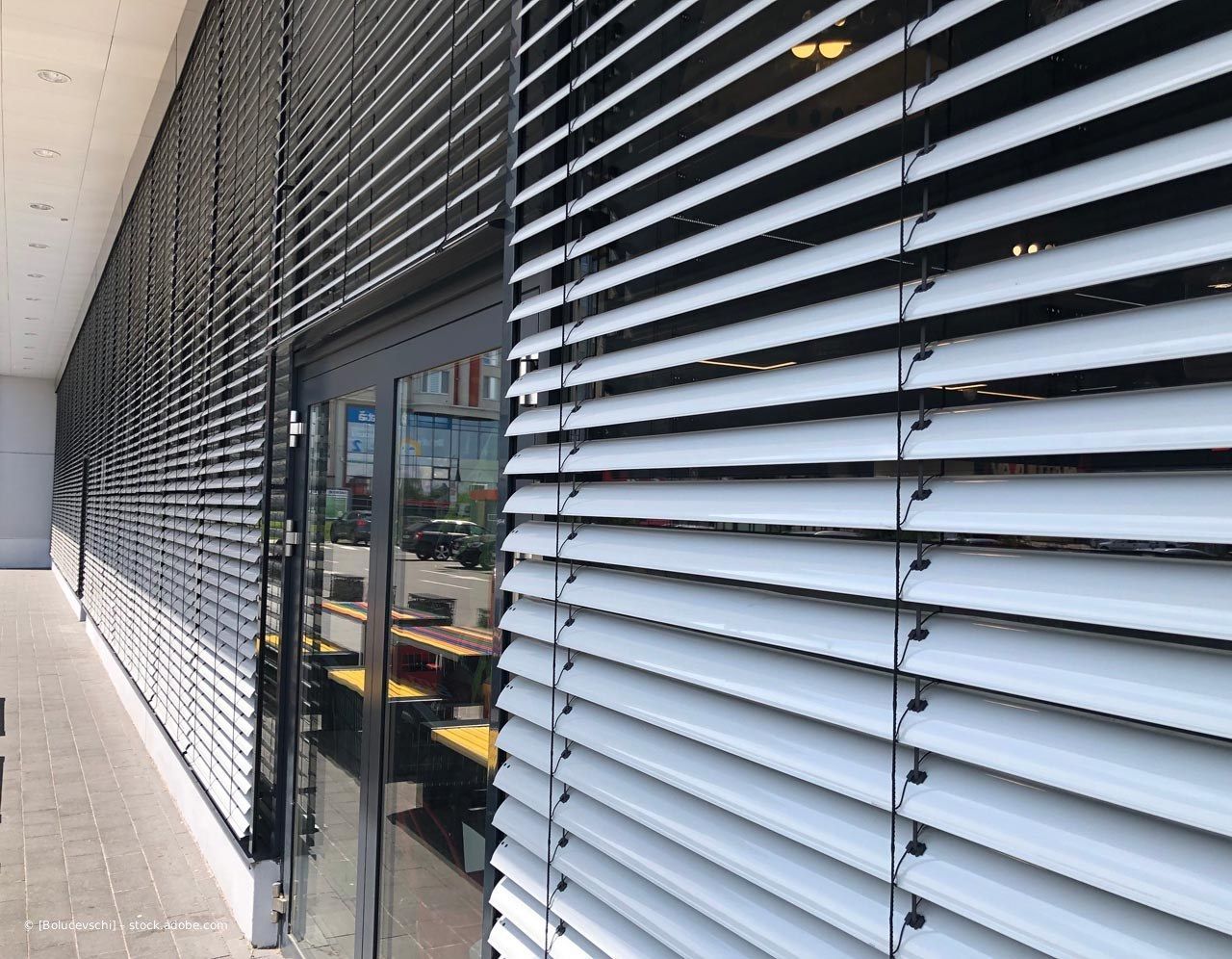 Außengehweg neben einer Glasfassade mit schwarzen, horizontalen Lamellen aus Metall.