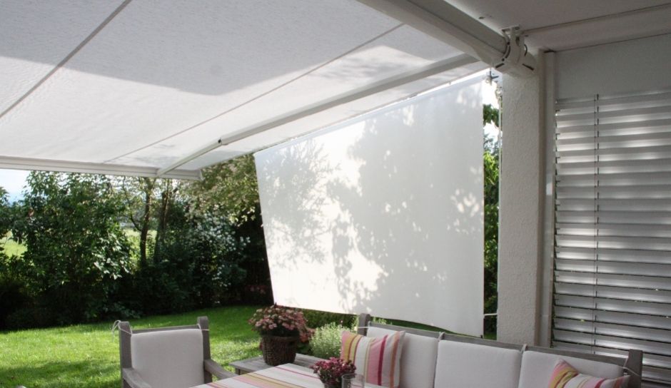 Überdachte Terrasse mit weißem, einziehbarem Sonnenschutz, Sitzgelegenheiten im Freien und dahinterliegendem grünen Rasen.