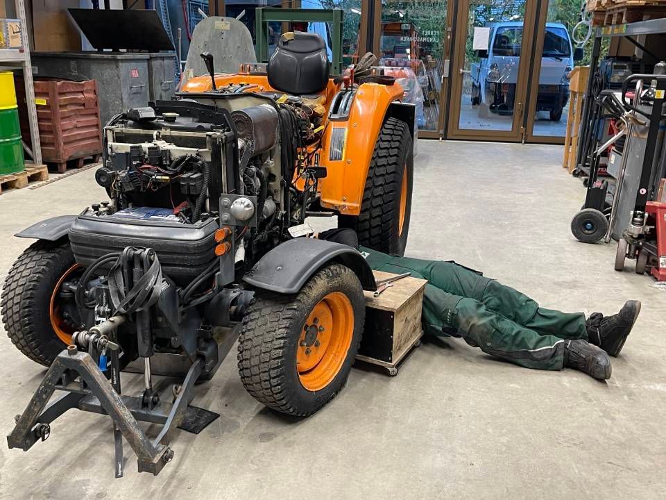 Ein Mechaniker arbeitet in einer Werkstatt an einem orangefarbenen Traktor und liegt dabei auf dem Boden.