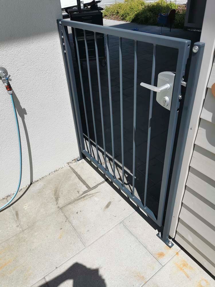 Ein Metalltor mit vertikalen Stäben und einem Griff, das in einem gepflasterten Bereich neben einer weißen Mauer steht.