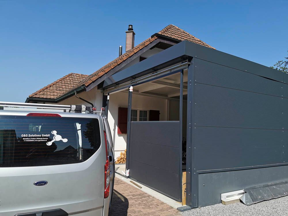 Ein grauer Carport an einem Haus, daneben parkt ein Lieferwagen. Strahlend sonniger Tag.