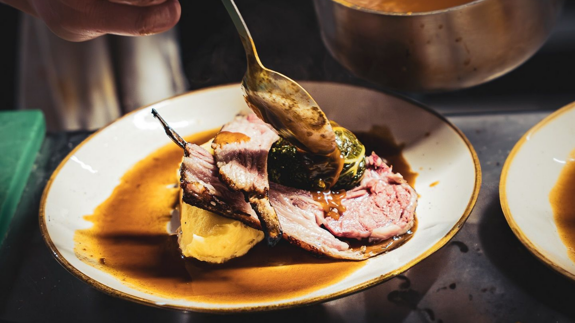 Une main utilise une cuillère pour verser une sauce brune sur de la viande rôtie et des légumes disposés dans une assiette