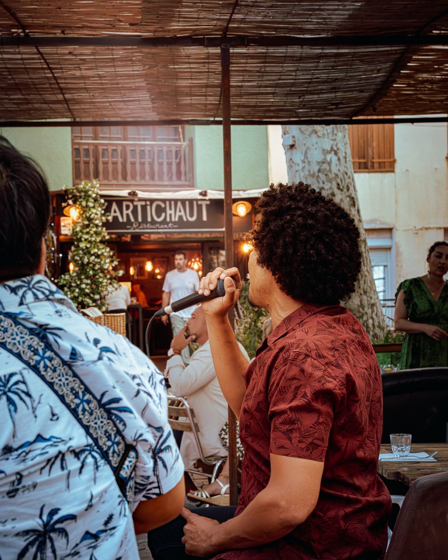 Une personne aux cheveux bouclés, vêtue d'une chemise rouge à motifs, chante dans un microphone lors d'un concert en plein air organisé à L'Artichaut