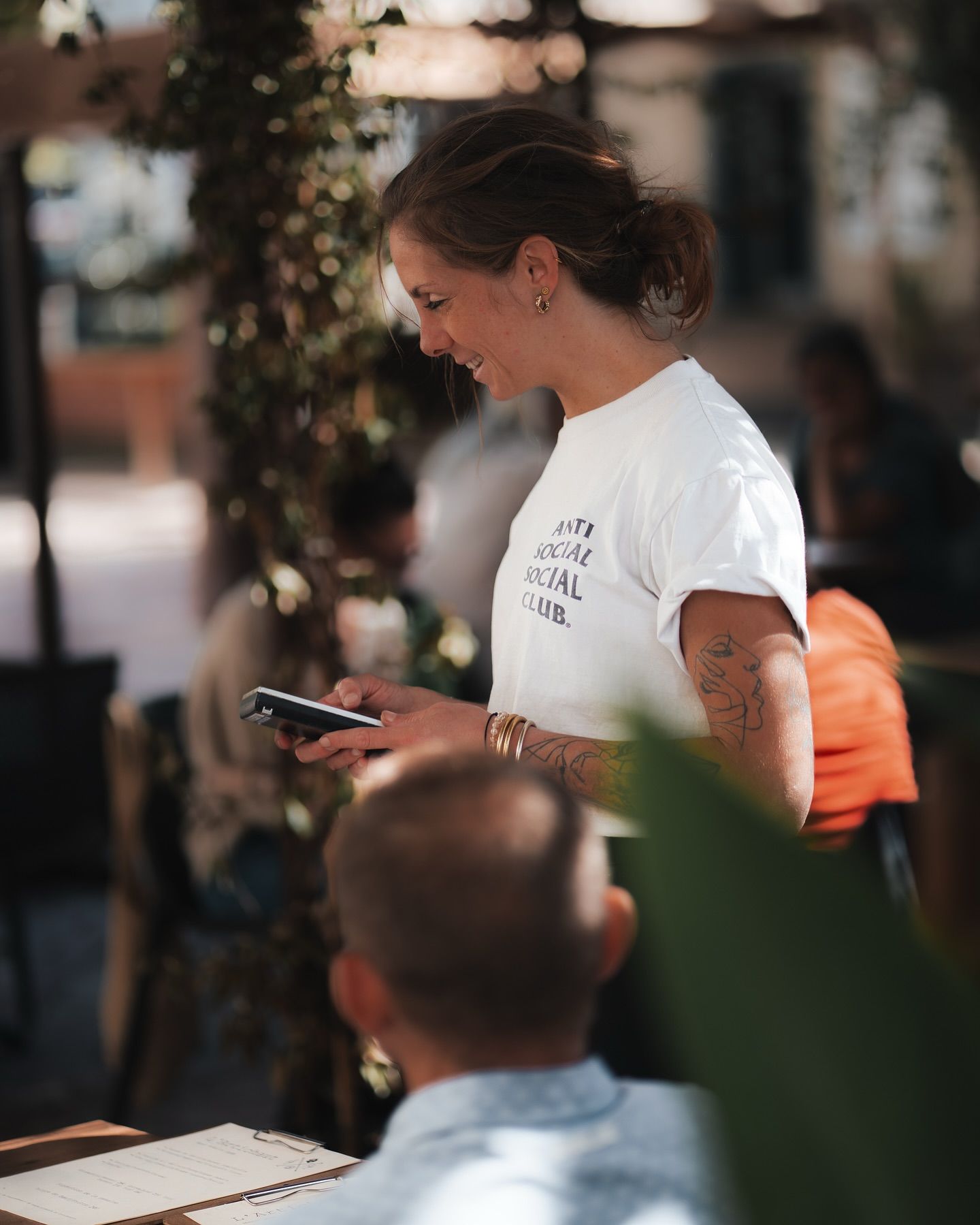 Une personne vêtue d'un t-shirt blanc sourit en regardant son smartphone dans un restaurant en terrasse