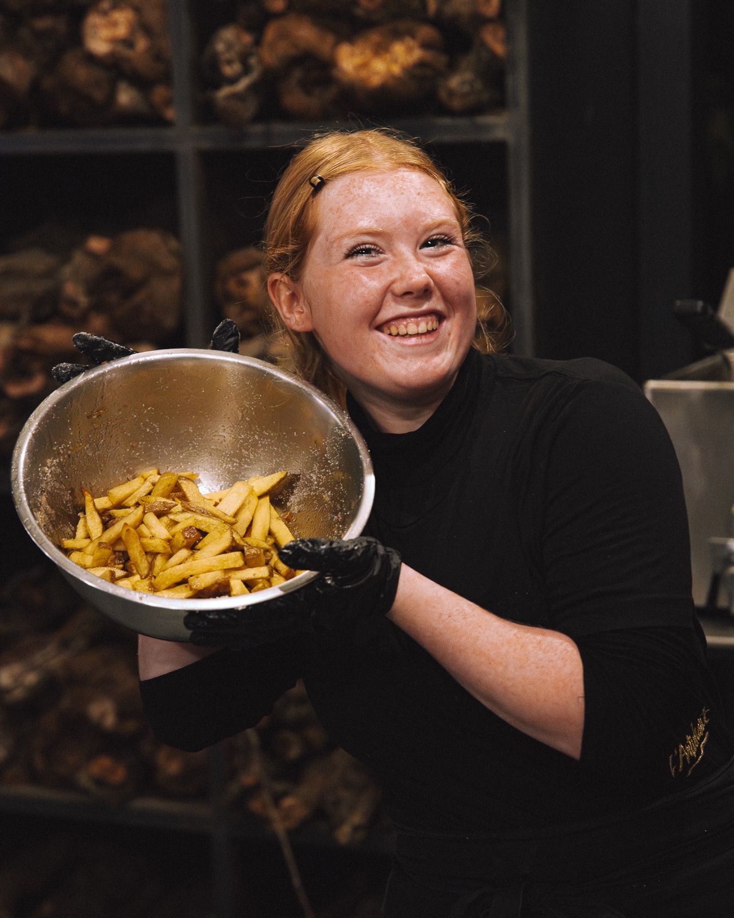 Dans une cuisine, une personne souriante, vêtue d'un uniforme noir et de gants, tient un grand bol en métal rempli de frites dorées